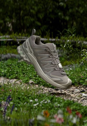 Chaussure de trail running Salomon marron clair avec dessus en mesh et semelle texturée flottant au-dessus de l'herbe et du sol rocailleux en extérieur.