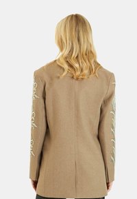 Blazer beige con tejido texturizado, que presenta intrincados detalles bordados en plata en ambas mangas y un corte entallado. Apto para ocasiones formales.