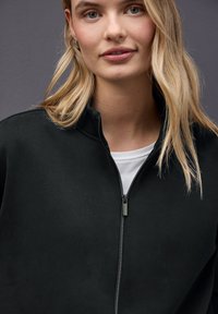 Chaqueta negra con cierre en la parte delantera y cuello alto, con un zipper plateado. Debajo, se asoma una camisa blanca. Textura suave, diseño minimalista.