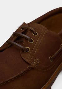 Καφέ δερμάτινο παπούτσι τύπου boat από σουέντ, με δερμάτινα κορδόνια περασμένα από ορειχάλκινα οπές και λεπτομερή ραφή.