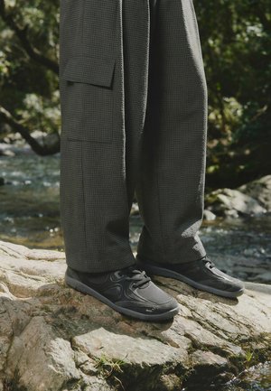 Osoba s nogama odjevena u široke tamne karirane hlače i crne tenisice stoji na kamenitoj površini blizu potoka u šumovitom području.