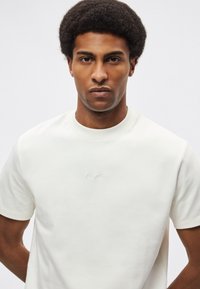 Krótka koszulka z krótkim rękawem w kolorze ecru, wykonana z gładkiej tkaniny, z minimalistycznym, haftowanym logo na piersi. Prosty krój z okrągłym dekoltem.