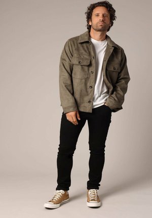 Veste en daim vert olive avec deux poches poitrine, t-shirt blanc, pantalon noir et baskets beige avec semelles blanches, debout sur un fond neutre.