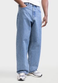 Άτομο που φοράει ανοιχτό μπλε τζιν παντελόνι με φαρδιά πόδια και αθλητικά παπούτσια ασημί-γκρι, με το ένα χέρι στην τσέπη και λευκό τοπ ορατό μερικώς.