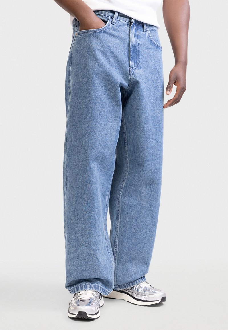 Άτομο που φοράει ανοιχτό μπλε τζιν παντελόνι με φαρδιά πόδια και αθλητικά παπούτσια ασημί-γκρι, με το ένα χέρι στην τσέπη και λευκό τοπ ορατό μερικώς.