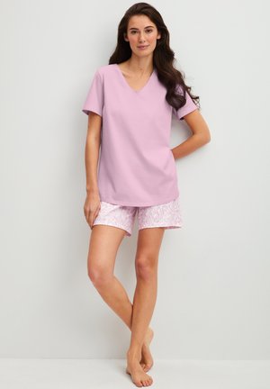 Vrouw staat blootsvoets tegen een lichte muur, draagt een lichtpaarse top met korte mouwen en witte shorts met roze bladpatronen.