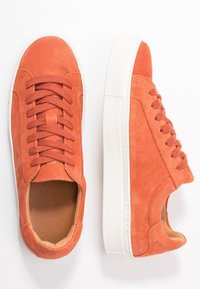 Oranžové semišové tenisky s bílými gumovými podrážkami, vybavené šesti očka, kulatými tkaničkami a minimalistickým designem. Vnitřek je z hnědé kůže.