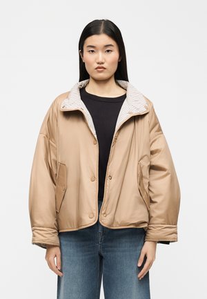 Donna che indossa una giacca oversize color tan con colletto interno a righe, maglietta nera e jeans blu, in piedi davanti a uno sfondo bianco e semplice.