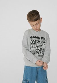 Šedá mikina s grafickým potlačem zobrazujícím herní ovladače. Volný střih, žebrované manžety a kulatý výstřih. Kombinováno s modrými cargo šortkami.