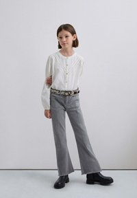 Blusa blanca de botones con mangas abullonadas, combinada con jeans acampanados grises y botas negras gruesas, acentuada por un cinturón estampado.