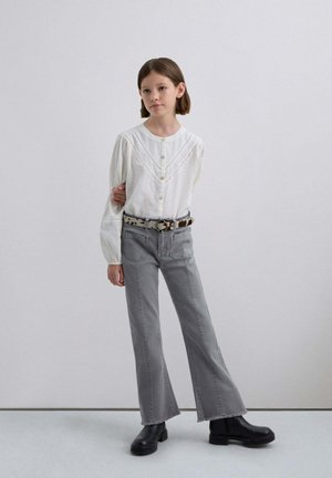 Blusa blanca de botones con mangas abullonadas, combinada con jeans acampanados grises y botas negras gruesas, acentuada por un cinturón estampado.