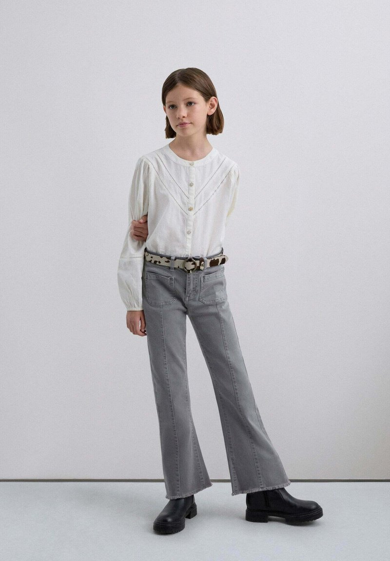 Blusa blanca de botones con mangas abullonadas, combinada con jeans acampanados grises y botas negras gruesas, acentuada por un cinturón estampado.