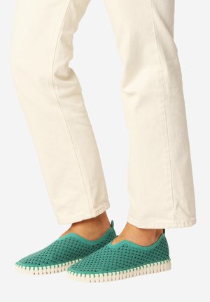 Para turkusowych butów wsuwanych z perforowanym wzorem noszonych z kremowymi spodniami, widocznych od połowy łydki w dół na jednolitym tle.