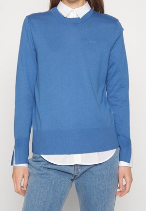 Maglione blu realizzato in materiale a maglia testurizzata, con scollatura rotonda, polsini a coste e un piccolo logo sul petto. Indossato sopra una camicia con colletto.