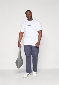 Weißes Baumwoll-T-Shirt mit dem Aufdruck "Originals Studio" in Schwarz, kombiniert mit marineblauen Hosen und hält eine graue Leder-Tasche. Neutraler Hintergrund.