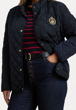 Femme portant une veste matelassée bleu marine avec un blason, un haut rayé rouge et bleu marine, un jean taille haute foncé et une ceinture noire avec une boucle dorée.