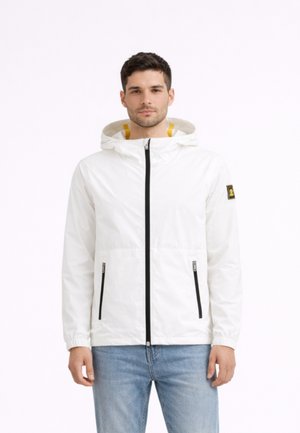 Uomo con giacca bianca con cappuccio, cerniera e tasche nere, in piedi davanti a uno sfondo bianco semplice.
