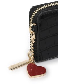 Portefeuille noir à motif crocodile avec fermeture éclair et accessoires dorés. Il présente un charme en forme de cœur rouge et une étiquette dorée attachée à la tirette de la fermeture éclair.