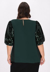 Blusa verde con maniche a palloncino in paillettes nere, dettaglio a oblò sulla schiena e corpo in tessuto morbido. Gli accenti testurizzati e lucidi sulle maniche contrastano con il corpo opaco.
