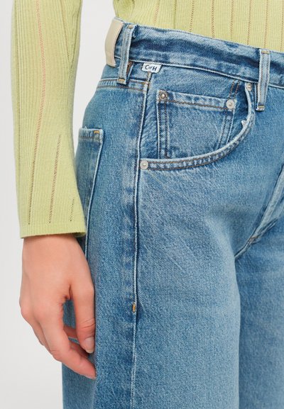 Jean en denim bleu clair à coupe décontractée, avec un design classique à cinq poches, des rivets en cuivre et une patch en cuir beige à la taille.