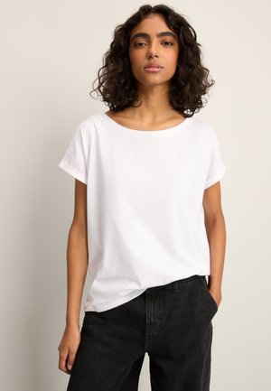 Femeie cu păr creț și închis la culoare, purtând un tricou alb simplu cu mâneci scurte și pantaloni negri, stând în picioare pe un fundal neutru.