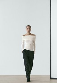 Kremowy top z opuszczonymi ramionami z teksturowanej tkaniny, w połączeniu z czarnymi, dopasowanymi spodniami. Prosty srebrny naszyjnik podkreśla linię dekoltu.