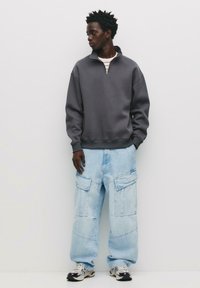 Graue Zip-Hoodie, hellblaue weite Cargo-Jeans und ein geschichtetes gestreiftes Hemd, ergänzt durch klobige Sneaker. Glatte Stofftextur.