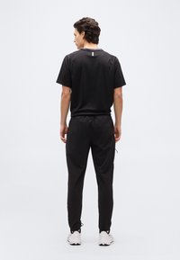 Νεαρός άνδρας στέκετα ι με την πλάτη γυρισμένη, φοράει μαύρο πουκάμισο με κοντά μανίκια, μαύρο παντελόνι και λευκά αθλητικά παπούτσια, μπροστά σε απλό φωτεινό φόντο.