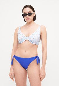 Bikini-set med lys blå rutetopp med rød kirsebærmønster og ensfarget blå bunn med knytedetaljer på sidene. Glatt tekstur hele veien.