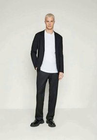 Μαύρο σακάκι πάνω από λευκό μπλουζάκι, συνδυασμένο με σκούρους παντελόνες και μαύρες γόβες. Ο ύφασμα φαίνεται λείος, με κομψή εφαρμογή.