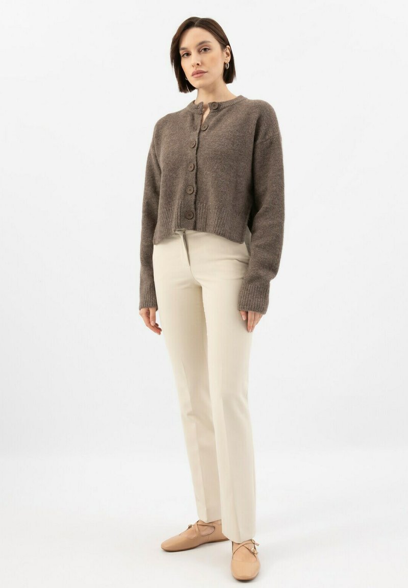 Hnědý zkrácený kardigan s texturovaným pletením a sedmi předními knoflíky, doplněný o smetanové upnuté kalhoty a béžové ploché boty.