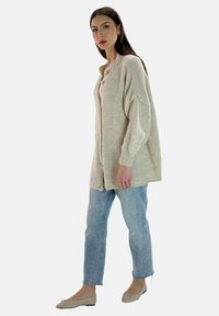 Beige stickad cardigan med knapplist framtill, nedsläppta axlar och strukturfinish, tillsammans med ljusblå denimjeans och beigefärgade ballerinas.