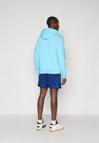 Sudadera azul claro con cordón, combinada con pantalones cortos azul oscuro que tienen un diseño de acento azul. Lleva puestos calcetines blancos y zapatillas beige.