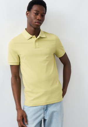 Jeune homme appuyé contre un mur blanc, portant un polo jaune clair et un jean bleu clair, avec une main dans la poche.