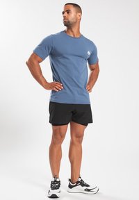 Heren blauwe katoenen T-shirt met korte mouwen, ronde hals en een klein wit logo op de borst, gecombineerd met zwarte sportieve shorts en schoenen.