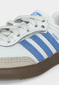 Κοντινή εικόνα λευκού sneaker Adidas Samba με μπλε ρίγες, λευκά κορδόνια και καφέ υφασμένη σόλα.