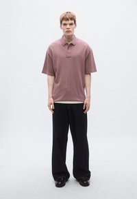 Πόλο μπλούζα κοντομάνικη σε μοβ χρώμα με υφή, με γιακά και λεπτομέρεια λογότυπου, συνδυασμένη με φαρδιά μαύρα τζιν παντελόνια και μαύρα παπούτσια.