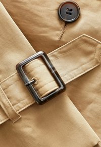 Tessuto beige con una texture liscia, caratterizzato da una grande fibbia rettangolare e un bottone scuro. Cuciture dettagliate.