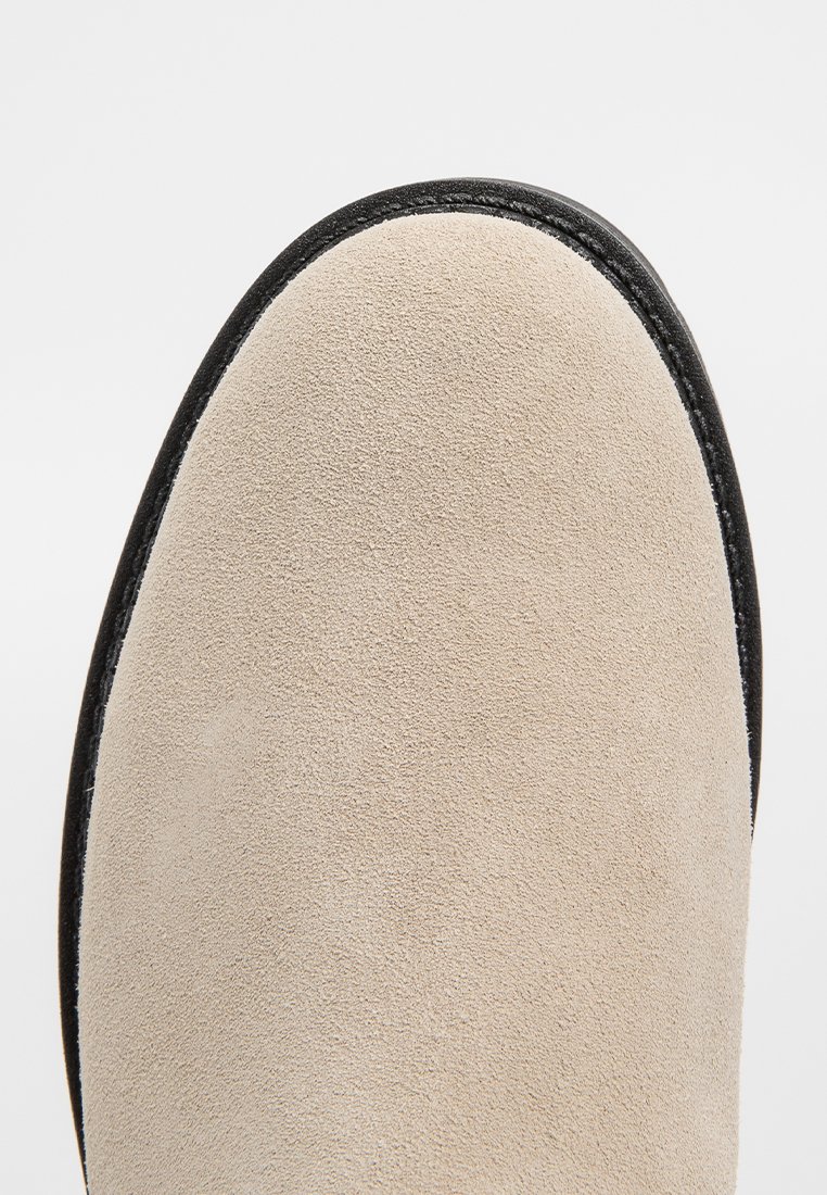 Beige Wildlederschuh mit runder Spitze und schwarzem Rand entlang der Kante. Die Oberfläche hat eine weiche Textur und ist für legere Anlässe konzipiert.