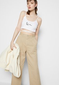 Weißes, kurzes Tanktop mit schwarzem Schriftzug-Logo, kombiniert mit hochtaillierten, beigen, weit geschnittenen Hosen. Leichte cremefarbene Jacke in der Hand.