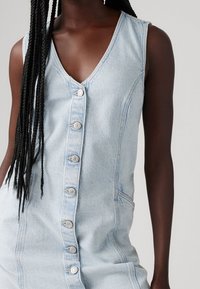 Mörkhyad person med långa flätor som bär en ärmlös ljusblå denimklänning med knappslå mot en enfärgad grå bakgrund.