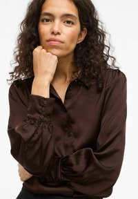 Blusa in satin marrone con chiusura a bottoni, maniche lunghe a sbuffo e colletto classico. Texture liscia con una leggera lucentezza e una vestibilità sagomata.