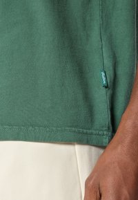 Πράσινο βαμβακερό t-shirt με ραμμένη γαζιά και πλαϊκή ετικέτα. Το μπλουζάκι έχει μαλακή υφή με λεία επιφάνεια. Η πλε�ιοψηφία της εικόνας δείχνει τον ύφασμα.