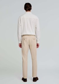 Chinos beige y una camisa blanca de botones con rayas vista desde atrás. Los chinos tienen bolsillos traseros con botones; la camisa presenta un cuello recto y puños.