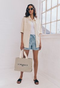 Camicia oversize color crema, top bianco, pantaloncini blu con fantasia, sandali neri e una borsa a righe con la scritta "LEMARIS". Ambientazione interna luminosa.