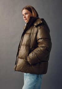 Puffjakke i mørk oliven grøn, med et quiltet design, hagebeskyttelse og oversized hætte. Har sidelommer og en tekstureret lynlås.