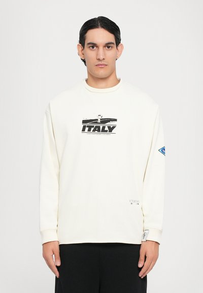 Crème oversized sweatshirt met een zwarte graphic met de tekst "ITALY." Inclusief rib gebreide manchetten en een patchdetail op de linker mouw.
