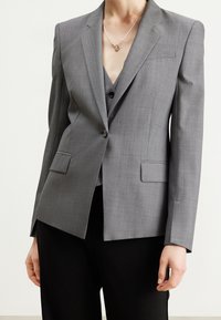 Blazer gris sur mesure avec un revers cranté, une fermeture à bouton unique, deux poches avant et des épaules structurées. Porté sur un gilet gris.