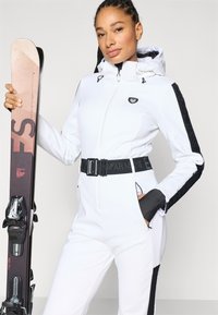 Λευκή ολόσωμη φόρμα σκι με μαύρα πλαϊνά πάνελ, ρυθμιζόμενη κουκούλα, ζώνη στη μέση και αθλητικό σχέδιο. Το άτομο κρατά ένα ζευγάρι σκι.