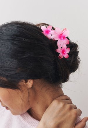 Pink plastic flower hair clip with three blossoms, positioned in a dark bun. Smooth texture with vivid colors and small holes for attachment.